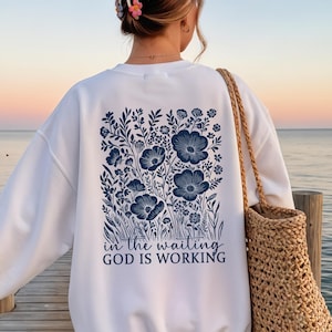 Könnte beinhalten: Weißes Sweatshirt mit marineblauem Blumendesign auf dem Rücken. Das Design umfasst Blumen, Blätter und den Text "in the waiting GOD IS WORKING". Eine Korbtasche wird über der Schulter getragen.