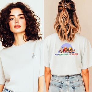 Könnte beinhalten: Hellgraues T-Shirt mit einer kleinen Handgesten-Grafik auf der Vorderseite und dem Text "MEIN STADTBILD IST DIVERS" mit einer Stadtszene auf der Rückseite. Das Shirt wird mit einer blauen Jeans getragen.