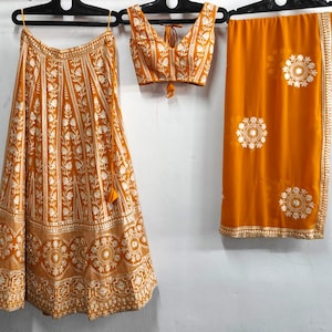 May include: A three-piece Indian ethnic wear set in a warm orange hue, featuring a skirt, a sleeveless top, and a draped shawl. The skirt and top are adorned with intricate white floral patterns. The shawl has white circular designs and a decorative border.