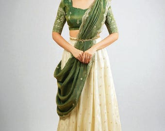 May include: A woman wearing a traditional Indian outfit. The ensemble includes a green, embroidered crop top with matching sleeves, a cream-colored skirt, and a draped green shawl. A gold belt cinches the waist, and the outfit is accessorized with earrings.
