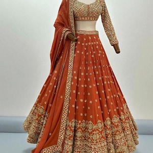 Puede incluir: Un conjunto lehenga naranja con intrincados bordados dorados. El conjunto incluye una falda larga, un top corto con mangas largas y una dupatta a juego. La falda y el top están adornados con patrones florales y geométricos.