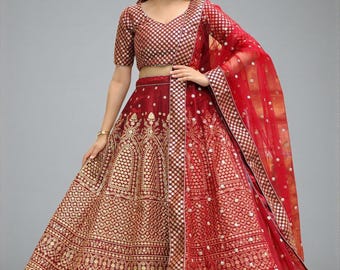 May include: A red and gold embroidered lehenga choli, a traditional Indian outfit. The ensemble features a short-sleeved top and a long skirt with intricate gold patterns. A matching red dupatta with gold trim completes the look. The outfit is suitable for festive occasions.