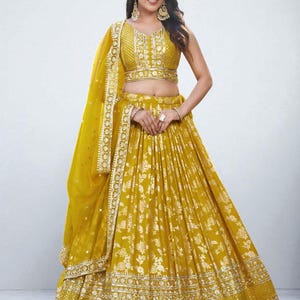 Puede incluir: Conjunto lehenga choli amarillo mostaza con bordado floral dorado. El top sin mangas y la falda presentan detalles intrincados, y el conjunto incluye un dupatta a juego. El atuendo está adornado con adornos dorados.