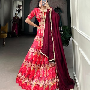 May include: A vibrant red and pink floral print lehenga with a matching cropped top and a burgundy dupatta. The lehenga features a flared skirt with a scalloped hemline. Gold trim accents the edges of the dupatta and top.
