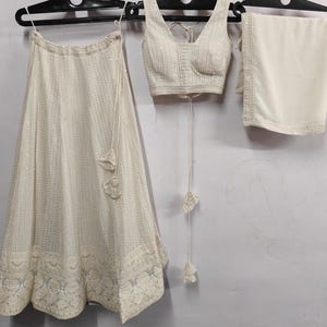 May include: Ivory-colored three-piece ethnic wear set. The set includes a skirt, a sleeveless top, and a shawl. The skirt has a detailed embroidered hem and a matching drawstring. The top has a V-neck and a matching drawstring. The shawl has a decorative border.