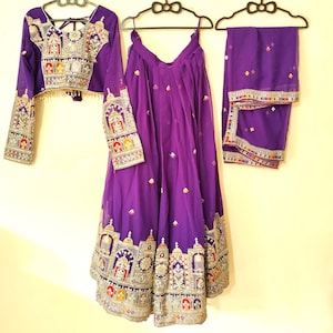 May include: A three-piece purple outfit with intricate embroidery. The set includes a long-sleeved top, a skirt, and a scarf. The embroidery features gold, red, and other colorful accents. The skirt and top have detailed patterns along the edges.