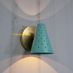 Peut inclure: Une applique murale avec une base circulaire de couleur laiton et un abat-jour conique. L'abat-jour est bleu clair avec des points noirs espacés. Le luminaire est monté sur un mur gris clair. Le texte "rihanhandcrafts" est visible.