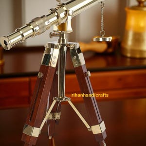 Puede incluir: Un telescopio de latón montado sobre un trípode de madera. El telescopio tiene un acabado metálico brillante y está conectado a un pequeño peso circular por una cadena. Las patas del trípode son de color marrón oscuro con detalles de latón. El texto "rihanhandicrafts" es visible.