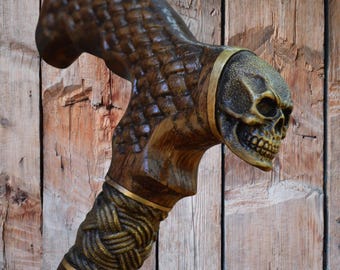 May include: A handcrafted wooden walking cane with a skull-shaped handle. The cane features intricate carvings and a detailed skull design. The handle is brown with gold accents, and the cane has a textured grip. The cane is a unique accessory.