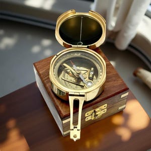 Puede incluir: Una brújula de latón vintage con tapa abatible, sobre una caja de madera. La esfera de la brújula muestra marcas direccionales y una aguja. La caja tiene un cierre de latón. La brújula está abierta, mostrando el interior.