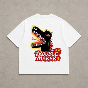 Pode incluir: Uma t-shirt branca de manga curta com um gráfico nas costas. O design mostra uma criatura preta com a boca aberta, dentes brancos afiados e uma língua vermelha. Abaixo da criatura, o texto TROUBLE MAKER está impresso em branco sobre um fundo vermelho.