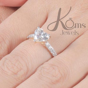 Könnte beinhalten: Ein herzförmiger Diamantring mit einem Band aus kleineren Steinen. Der Ring befindet sich an einer Hand mit manikürten Nägeln. Der Markenname "Koms Jewels" ist im Hintergrund sichtbar.