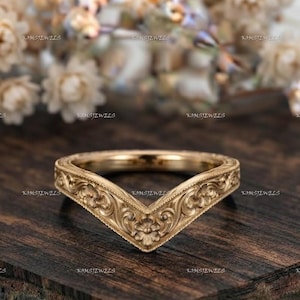 May include: A V-shaped gold ring featuring an intricate engraved floral scroll pattern across the entire outer band. The metal has a warm, polished finish, and the design forms a distinct chevron point at the center of the band's circumference.