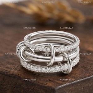 Può includere: Un anello multi-fascia color argento con due fasce esterne incastonate di piccole pietre chiare e una fascia centrale liscia e lucida. Tre piccoli anelli color argento sono intrecciati attraverso la struttura principale.