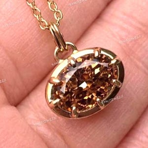 以下が含まれることがあります： 金色のネックレスと楕円形のペンダント。ペンダントは、金色のベゼルにセットされた、大きくファセットカットされた茶色の宝石が特徴です。チェーンとペンダントループも金色です。背景はぼやけた手です。