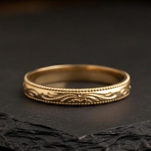 Puede incluir: Un anillo dorado con un intrincado patrón repetitivo alrededor de la banda. El anillo se encuentra sobre un fondo oscuro, resaltando su diseño detallado. El anillo parece ser de metal y probablemente sea una alianza de boda o una joya similar. Diámetro aproximado de 2 cm.