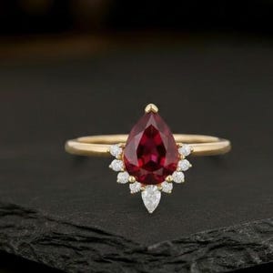 May include: A gold ring featuring a large, pear-shaped, deep red gemstone as the focal point. The gemstone is surrounded by smaller, clear accent stones. The ring is set on a black surface.