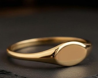 May include: A gold signet ring with a polished finish. The ring features a smooth, oval-shaped face and a slender band. The ring is displayed on a dark surface, with a blurred background.