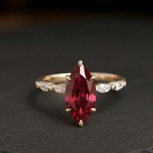 May include: A gold ring featuring a marquise-cut, deep red gemstone as the centerpiece. The band is adorned with smaller, clear gemstones. The ring is set against a dark background, highlighting its elegant design.