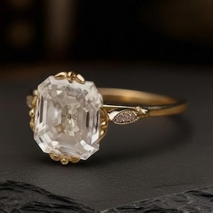 May include: A gold ring featuring a large, clear, emerald-cut gemstone. The ring has a delicate band and ornate detailing around the gemstone setting. Small accent stones are set on the band near the gemstone.