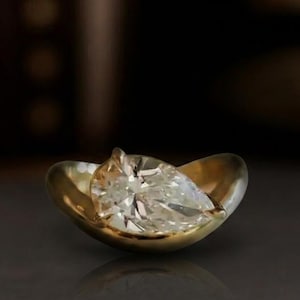 May include: A pear-shaped diamond ring set in a gold, petal-like setting. The diamond sparkles with many facets, reflecting light. The ring is displayed on a dark, textured surface, showcasing the jewelry's brilliance.