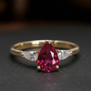 Puede incluir: Anillo de oro con una piedra roja en forma de pera en el centro, flanqueada por dos piedras claras más pequeñas. La banda del anillo es dorada y el engaste es delicado. Joyería para mujer.