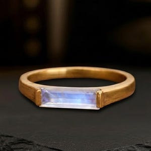 May include: A gold ring featuring a single, horizontal, rectangular baguette-cut moonstone. The band has a smooth, polished finish, and the gemstone displays a soft, iridescent blue glow, set securely within the metal frame of the ring.