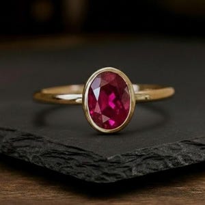 Könnte beinhalten: Ein goldener Ring mit einem großen, oval geschliffenen Rubin-Edelstein. Der Rubin hat eine tiefrote Farbe und ist in einer goldenen Lünette gefasst. Der Ring wird auf einer schwarzen Oberfläche präsentiert.