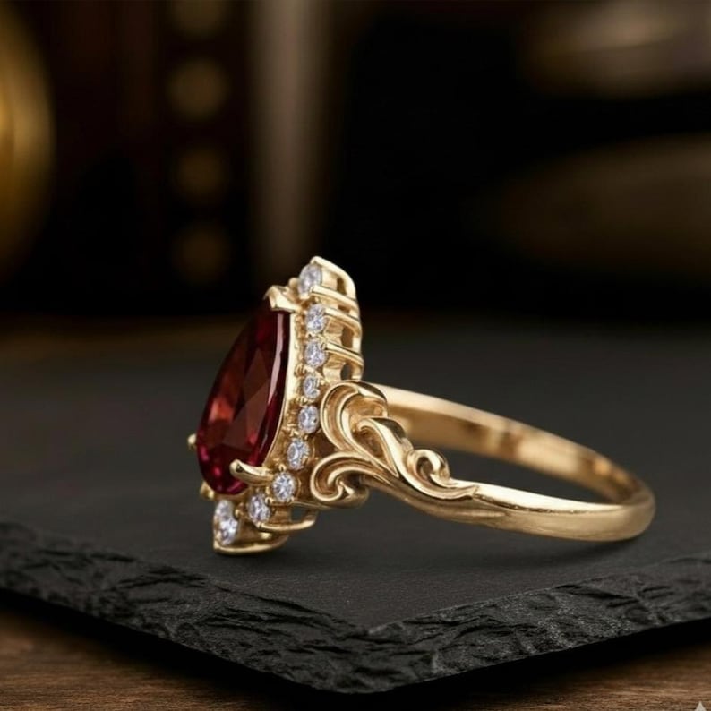 May include: A gold ring featuring a teardrop-shaped, deep red gemstone. The ring is embellished with small, clear accent stones and has an elaborate, swirling pattern on the band.