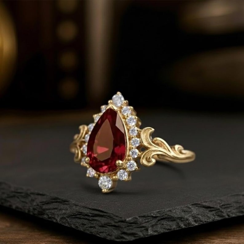 May include: A gold ring with a large, teardrop-shaped, deep red gemstone, encircled by smaller, clear stones. The ring has an elaborate, swirling design on the band and around the central stone. The ring is shown on a black surface.