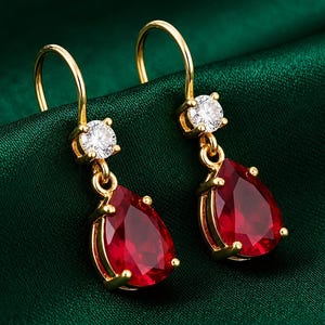 May include: A pair of gold drop earrings featuring a pear-shaped red gemstone suspended from a small, round, clear white stone. The earrings are designed with a curved hook closure, showcasing a polished gold setting that secures each vibrant red teardrop-shaped stone with four prongs.