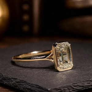 Puede incluir: Un anillo de oro con una gema grande, rectangular y transparente. El anillo está sobre una superficie oscura y texturizada. La banda y el engaste son de color oro brillante, creando un contraste llamativo con la piedra.