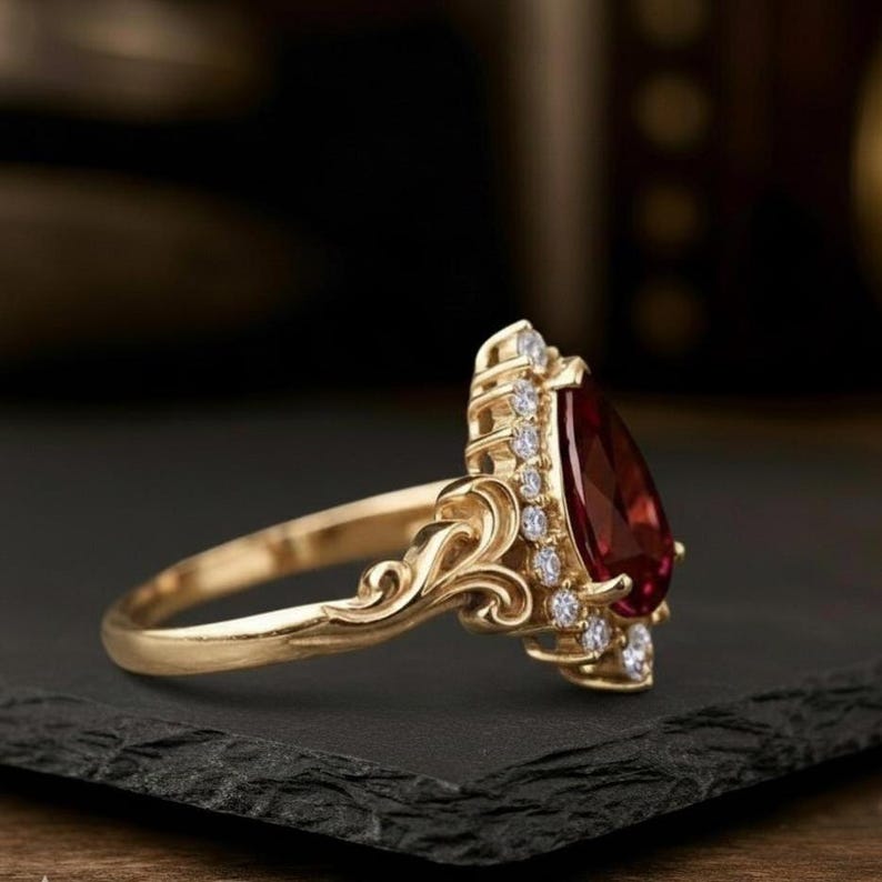May include: A gold ring with a teardrop-shaped, deep red gemstone, surrounded by small, clear gemstones. The ring has an ornate, filigree design on the band and around the central stone, giving it a vintage appearance.