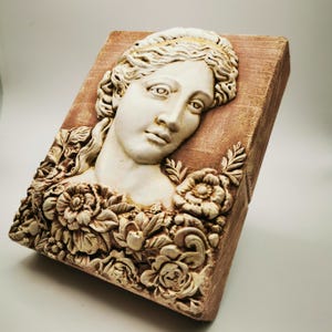 Könnte beinhalten: Rechteckige Reliefskulptur mit einem Frauengesicht, umgeben von floralen Details. Die Skulptur ist cremefarben und vor einem strukturierten, rotbraunen Hintergrund platziert. Das Gesicht der Frau ist detailliert, mit fließendem Haar und einem ruhigen Ausdruck. Wahrscheinlich eine Wanddekoration.