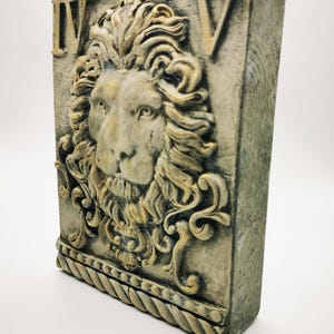 Könnte beinhalten: Rechteckige, verwittert aussehende Wandplakette mit einem detaillierten Relief eines Löwenkopfes mit fließender Mähne. Die Plakette hat die römischen Ziffern IV und V in den oberen Ecken. Das Löwengesicht ist hellbeige, und die Plakette hat einen seilartigen Rand.
