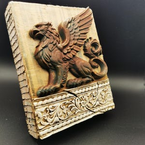 Könnte beinhalten: Antik anmutende Wandplakette mit einem Relief eines Greifen. Der Greif, in Bronzetönen, ist vor einem beigen Hintergrund platziert. Die Plakette hat einen weißen, verzierten Rand und einen Riss-Effekt. Maße: ca. 20 cm x 15 cm.