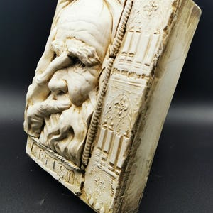 Könnte beinhalten: Beige rechteckige Reliefskulptur mit einem detaillierten Profil eines bärtigen Mannes. Die Skulptur trägt das Wort "ARTIST" am unteren Rand und einen strukturierten Rand. Die Seite der Skulptur hat ein geschnitztes Gebäudedesign.