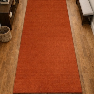 May include: A long, rectangular runner rug in a solid, vibrant burnt orange color. The rug features a consistent, low-pile texture and is positioned centrally on a wooden floor, extending lengthwise through the middle of the hallway space.