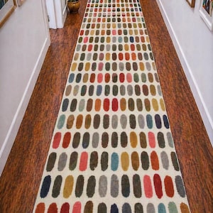 Puede incluir: Una alfombra larga y rectangular con un fondo crema y un patrón repetitivo de formas ovaladas coloridas. Los óvalos son en tonos de rojo, rosa, marrón, azul y gris. La alfombra está colocada en un pasillo con suelo de madera.