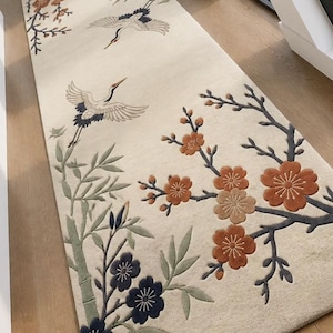 Op de afbeelding: Een rechthoekig, crèmekleurig tapijt met een oosters ontwerp. Het tapijt heeft geborduurde afbeeldingen van kraanvogels, bamboe en bloeiende takken in bruin-, groen- en marineblauw. Het tapijt ligt op een houten vloer.