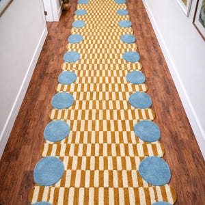 May include: A long hallway rug featuring a repeating pattern of mustard yellow and cream-coloured rectangles. Light blue, circular accents are placed along the edges. The rug is placed on a dark wood floor.
