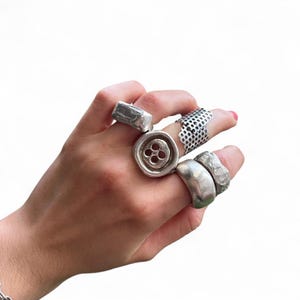 May include: A hand adorned with several silver rings. The rings showcase diverse designs, such as a button-style ring, a perforated band, and rings with textured surfaces. The rings are presented against a white backdrop.