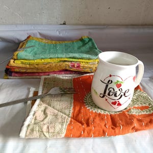Puede incluir: Una taza de cer&aacute;mica blanca con la palabra "Love" en escritura, decorada con corazones rojos, se encuentra sobre un mantel individual naranja y verde estampado. Tambi&eacute;n se ve una pila de cuadrados de tela cosidos de colores y un tenedor.