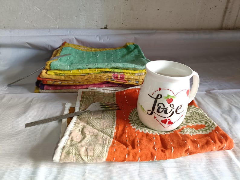 Puede incluir: Una taza de cer&aacute;mica blanca con la palabra "Love" en escritura, decorada con corazones rojos, se encuentra sobre un mantel individual naranja y verde estampado. Tambi&eacute;n se ve una pila de cuadrados de tela cosidos de colores y un tenedor.
