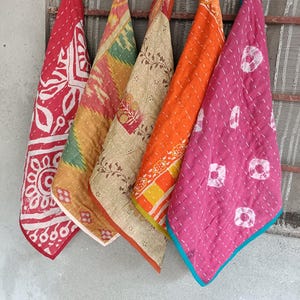 Könnte beinhalten: Fünf farbenfrohe, gesteppte Stoffstücke hängen vor einem Fenster. Die Stoffe sind rot, gelb, orange und rosa, mit verschiedenen Mustern, darunter Blumen- und geometrische Designs. Jedes Stück hat ein einzigartiges Design und ist mit weißem Faden genäht.