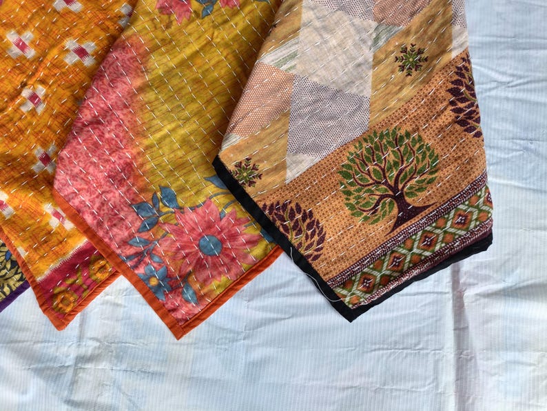 Pode incluir: Close-up de tr&ecirc;s t&ecirc;xteis coloridos estampados. Os tecidos apresentam desenhos florais e geom&eacute;tricos em tons de laranja, rosa, amarelo e castanho. As bordas s&atilde;o finalizadas com uma borda contrastante e costuradas com fio branco. Os t&ecirc;xteis parecem ser feitos &agrave; m&atilde;o.