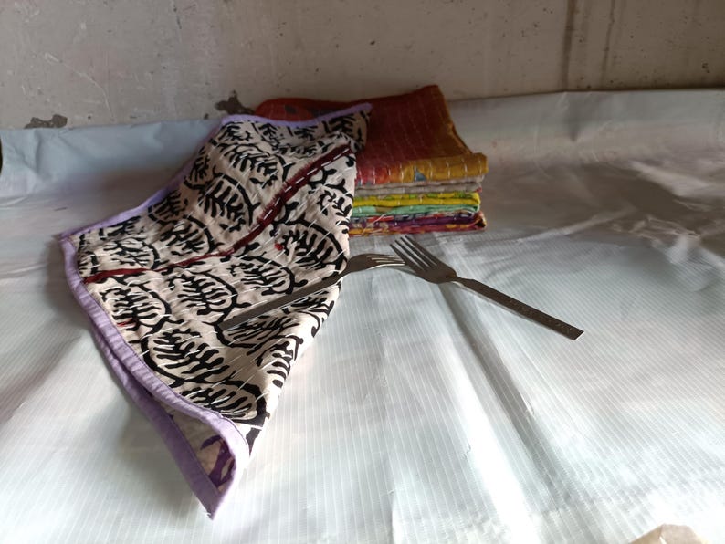 Pode incluir: Uma toalha de tecido dobrada creme com um padr&atilde;o de folhas pretas e uma borda roxa. Uma pilha de guardanapos coloridos est&aacute; ao fundo. Dois garfos prateados est&atilde;o colocados na superf&iacute;cie branca.