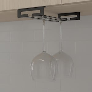 May include: A black metal wine glass rack fixed under a cabinet, holding two clear wine glasses. The glasses are suspended upside down, showing their elegant design. The background features a white tiled wall.