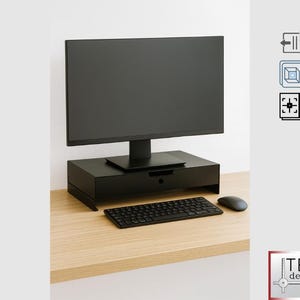 May include: A black computer monitor is positioned above a black monitor stand with a drawer. A black keyboard and mouse are on the light wood desk. The image also includes a logo that says "TEC design".