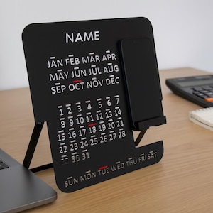 May include: A black desk calendar with a phone holder. The calendar displays the months and dates, with the word "NAME" at the top. A laptop, calculator, notepad, and pen are also on the desk.